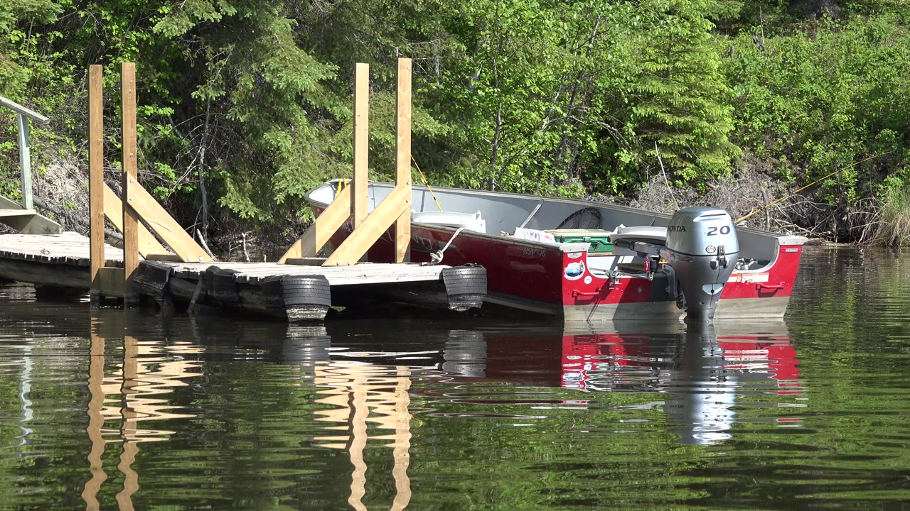 A small red and gray motorboat is tied beside a wooden dock in Red Lake, Canada, with black dock bumpers, green trees behind it, and no visible people.