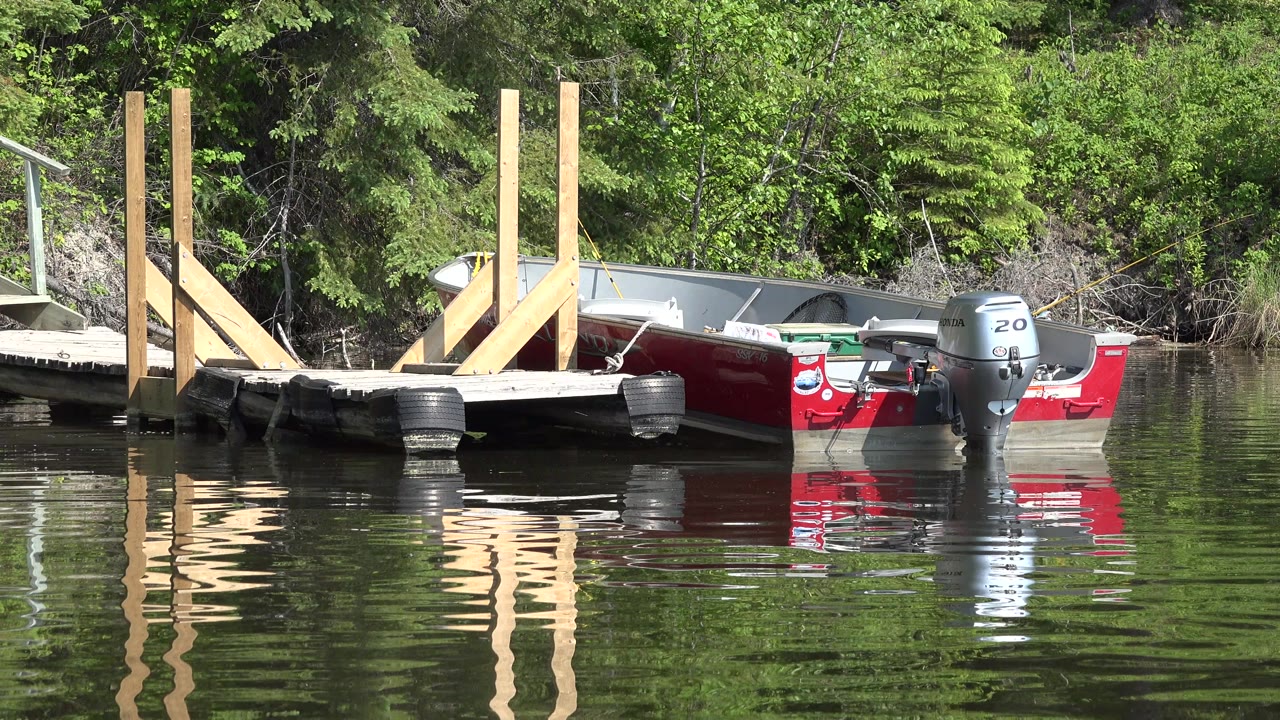 The boat remains beside the dock in Red Lake, Canada, while the dock posts and red hull reflect in a slightly rippled water surface with no visible people.