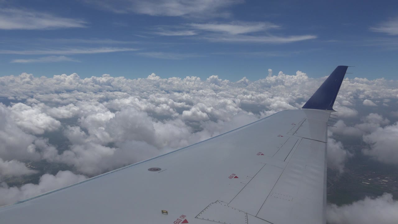 At 0:00, a gray airplane wing and blue-tipped winglet stretch over cloud layers above Winnipeg, Canada, with patches of ground below and no visible people.