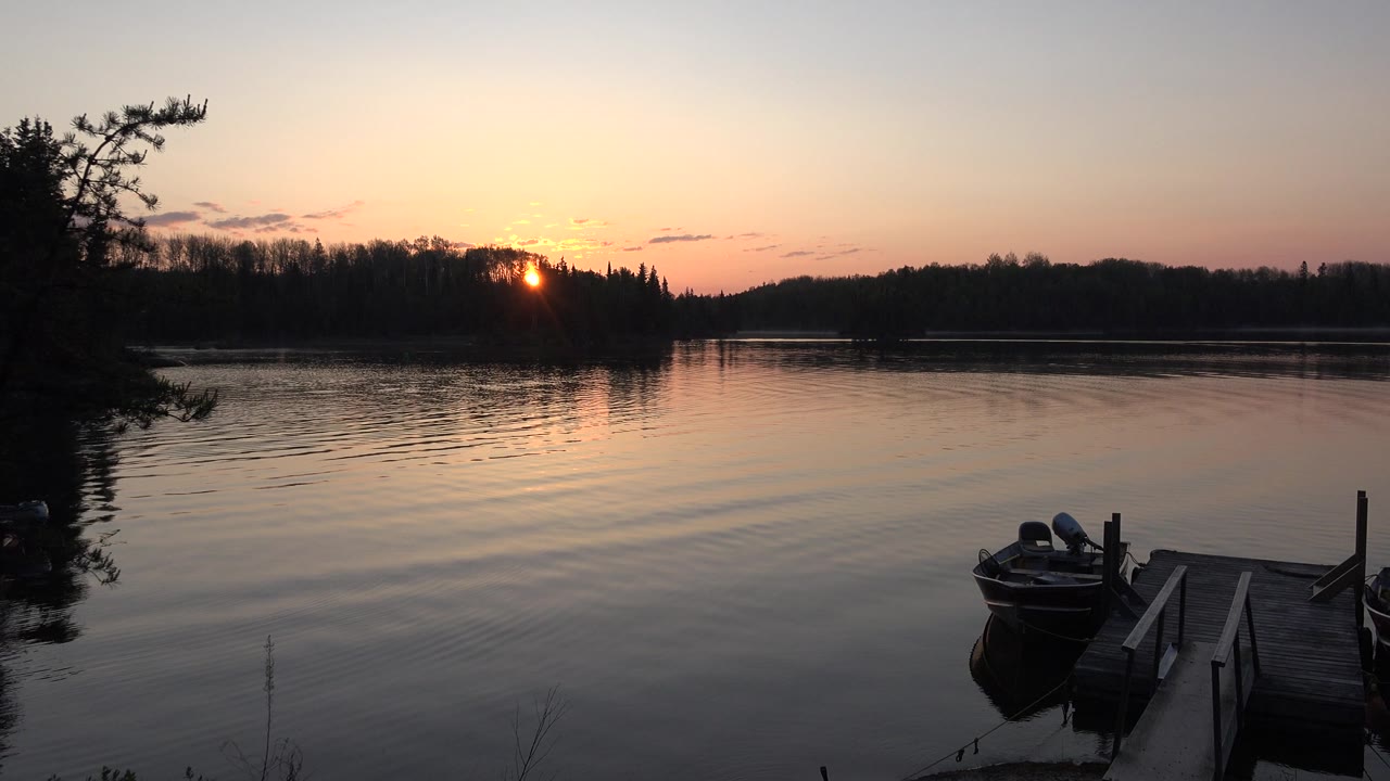 Red Lake, Canada sunset view with the sun partly hidden by the tree line, a narrow reflection on the water, the docked boat at lower right, and no visible people.