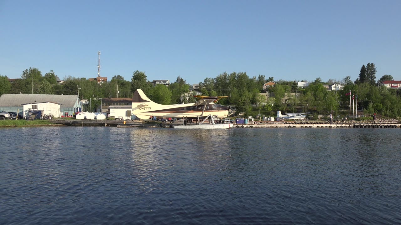 Wide view across rippled lake water toward a Red Lake, Canada shoreline with a cream-colored seaplane, low buildings, trees, and no visible people.
