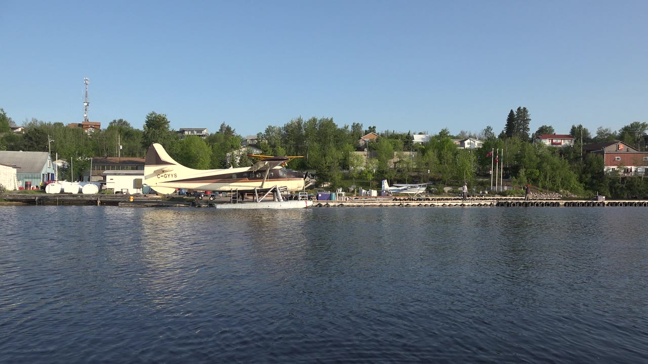 The Red Lake, Canada shoreline remains in a wide composition, with the seaplane near the docks, green trees behind the buildings, rippled water, and no visible people.