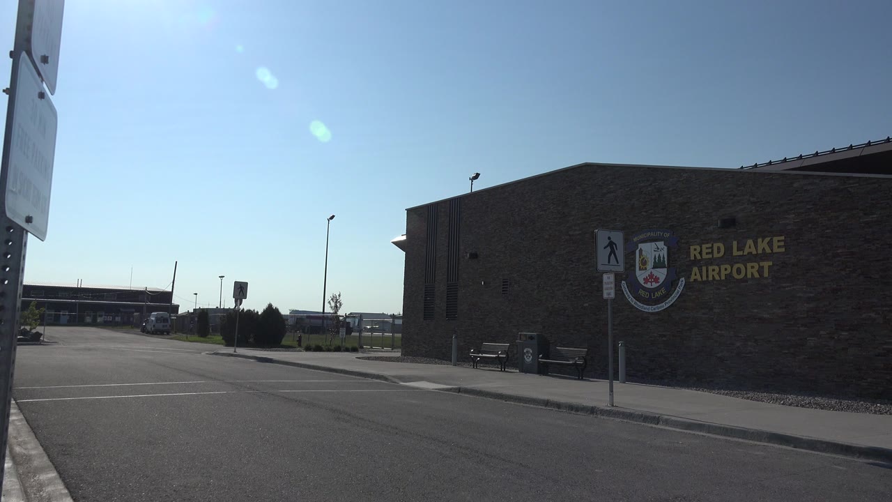 Opening view of the Red Lake Airport exterior in Red Lake, Canada, with the stone wall, yellow airport lettering, pedestrian sign, asphalt curb, blue sky, lens flare, and no visible people.
