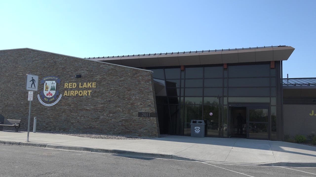 Red Lake, Canada airport exterior at the start of the clip, showing the stone wall, Red Lake Airport sign, glass entry, sidewalk, curb, and no visible people.