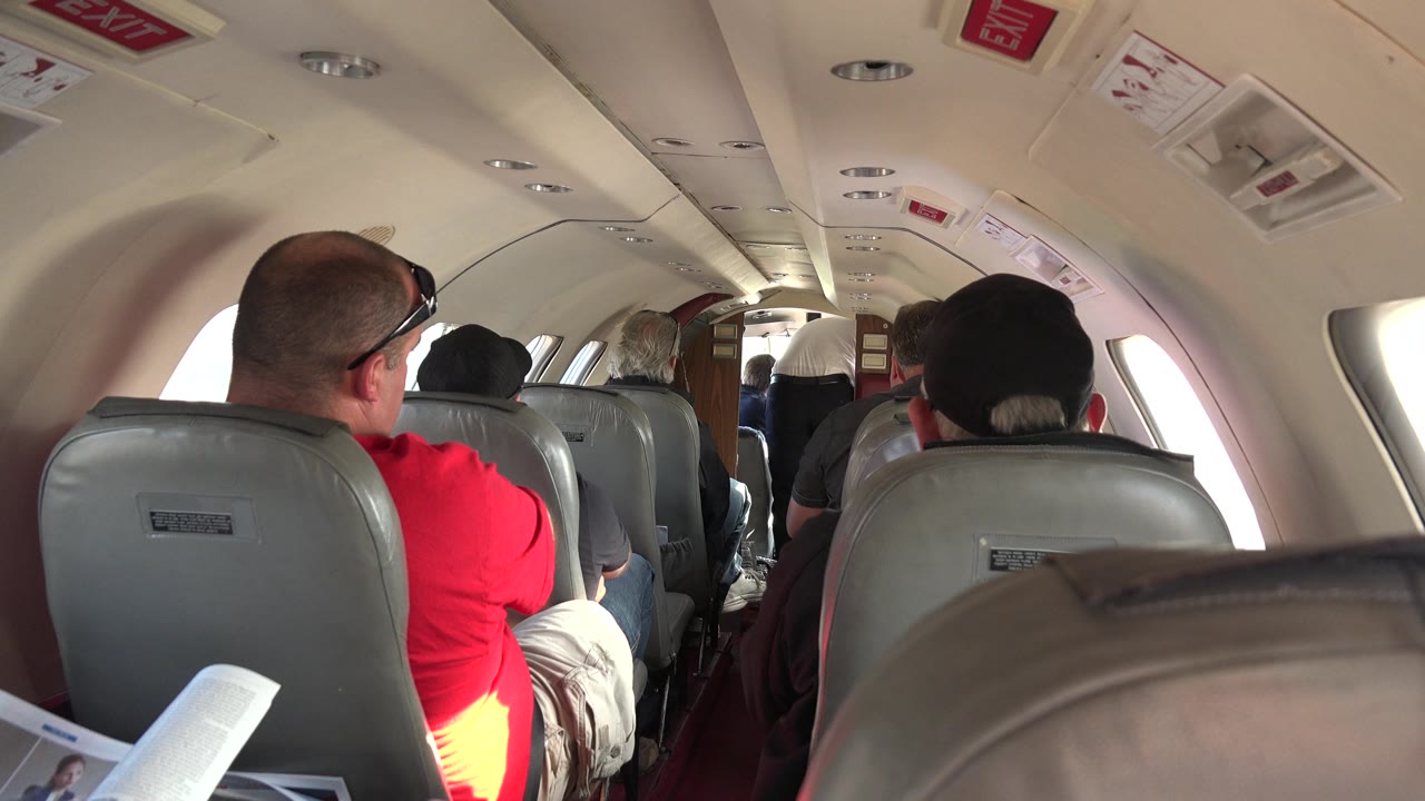 Passenger-seat perspective inside the Red Lake, Canada flight, showing a close tan seatback, gray rows ahead, and adults seated under overhead safety placards.