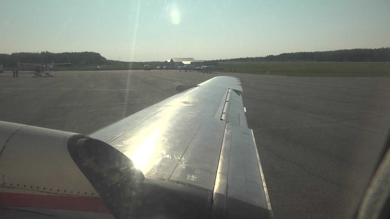 At 0:00 in Red Lake, Canada, a silver airplane wing fills the foreground while small aircraft sit beyond the paved apron under a hazy sky, with no visible people.