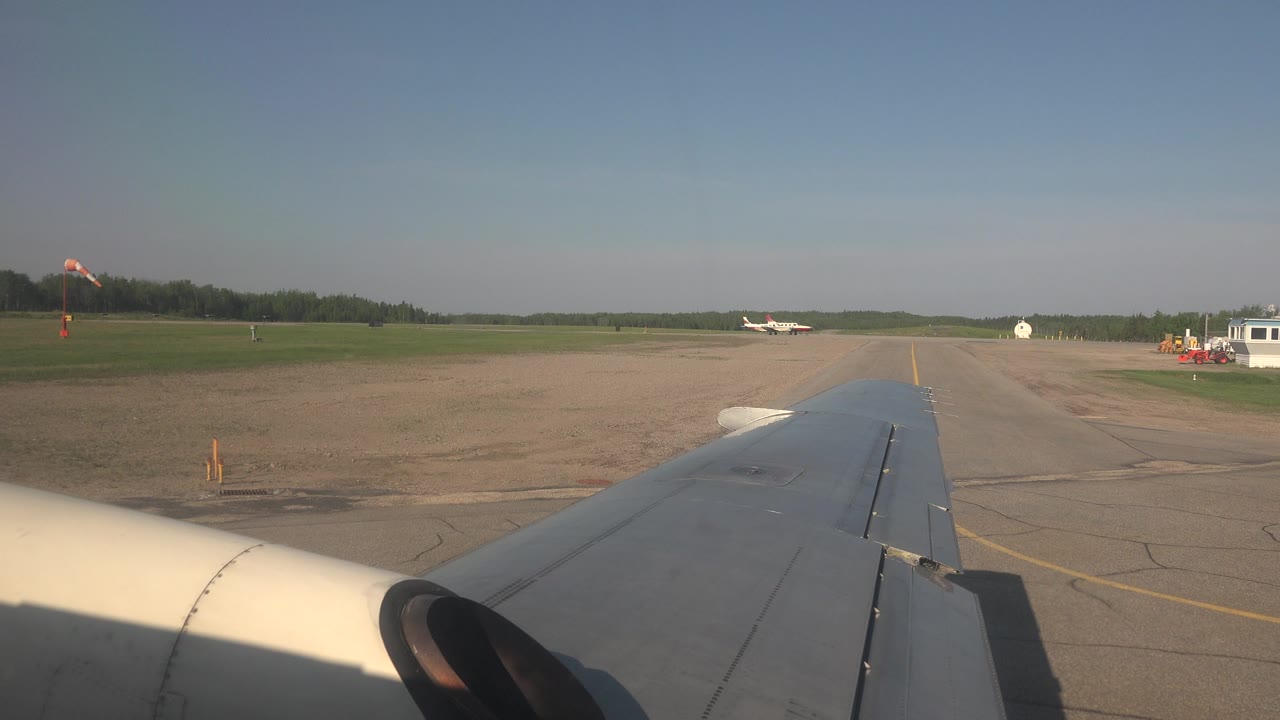 Passenger-window view in Red Lake, Canada, showing the airplane wing over cracked pavement, a small aircraft in the distance, a windsock, grass, trees, and no visible people.