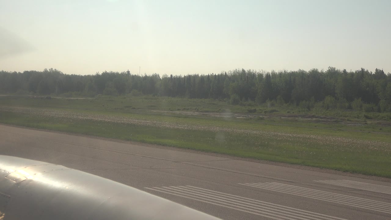 Aircraft metal in foreground beside a runway and treeline.