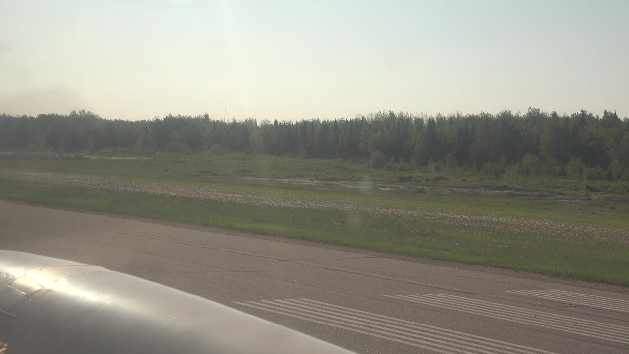 The aircraft foreground catches bright light while runway lines, a green field, shallow water patches, and trees remain visible at Red Lake, Canada, with no visible people.