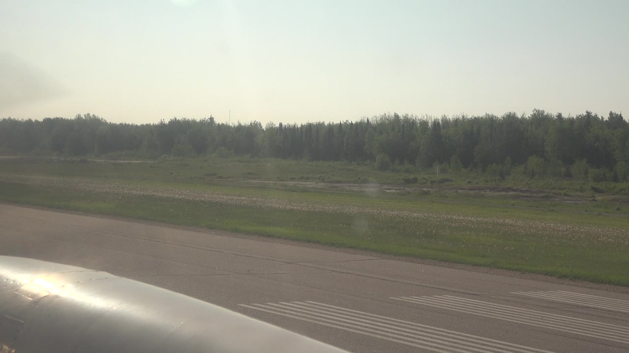 At 0:00 in Red Lake, Canada, a shiny airplane foreground sits along the bottom edge with runway markings, green field, distant trees, hazy sky, and no visible people.