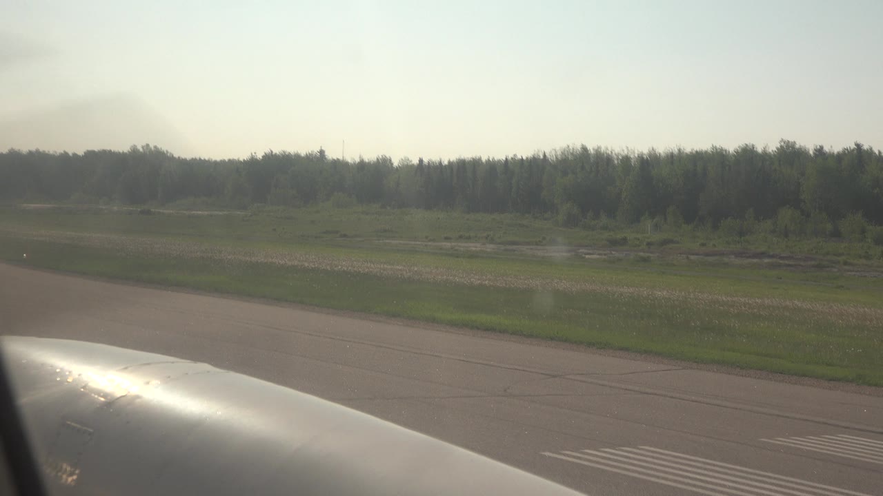 Passenger-window view in Red Lake, Canada, showing a bright airplane surface at the lower edge, runway pavement, green grass, distant forest, and no visible people.