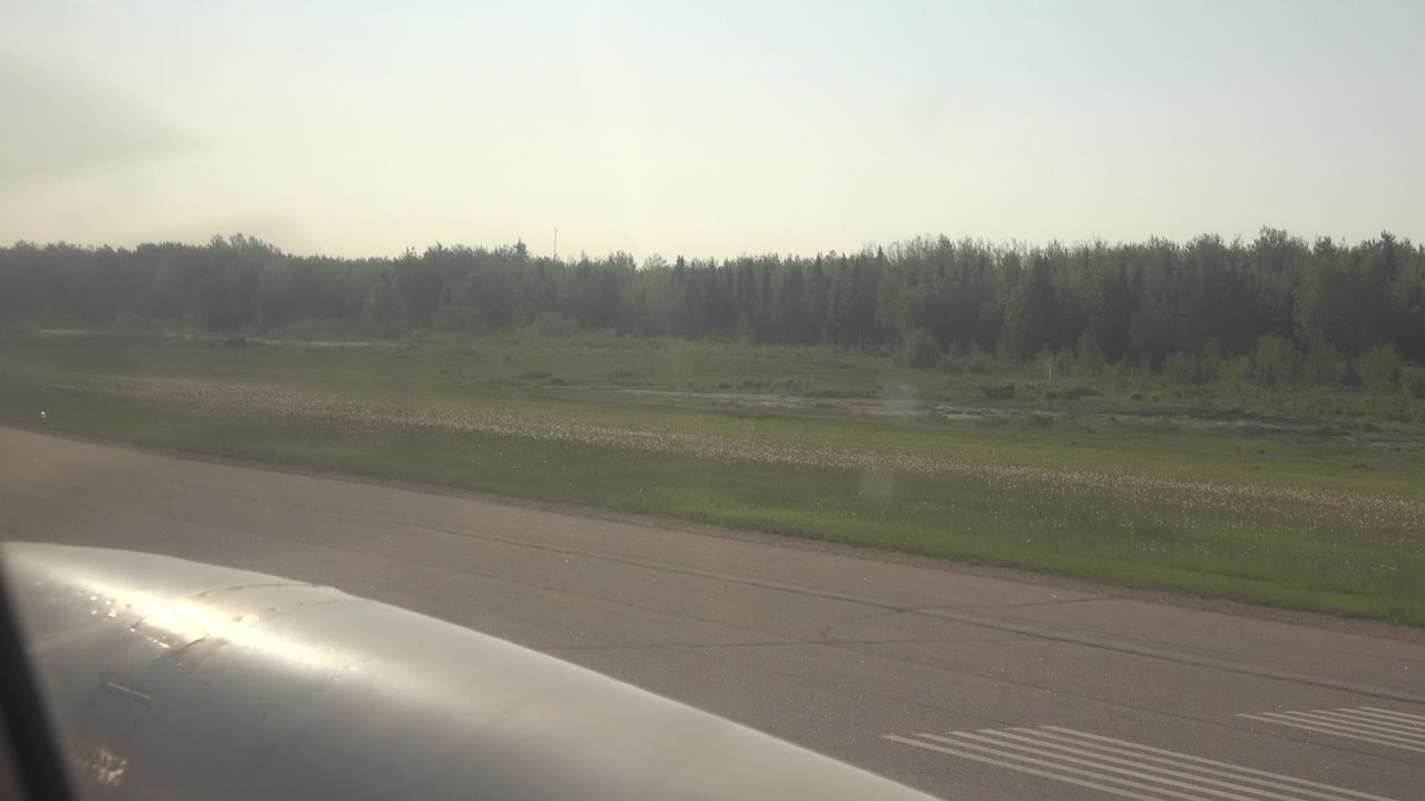 Second frame from the passenger window in Red Lake, Canada, with the airplane surface catching sunlight, runway markings below, forest in the distance, and no visible people.
