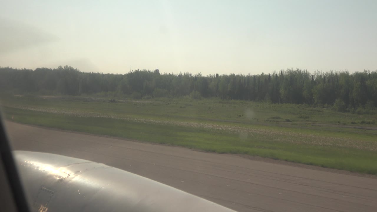 Second frame in Red Lake, Canada with pavement in front, green field across the middle, a distant wooded horizon, and a dark vertical aircraft window edge near the foreground with no visible people.