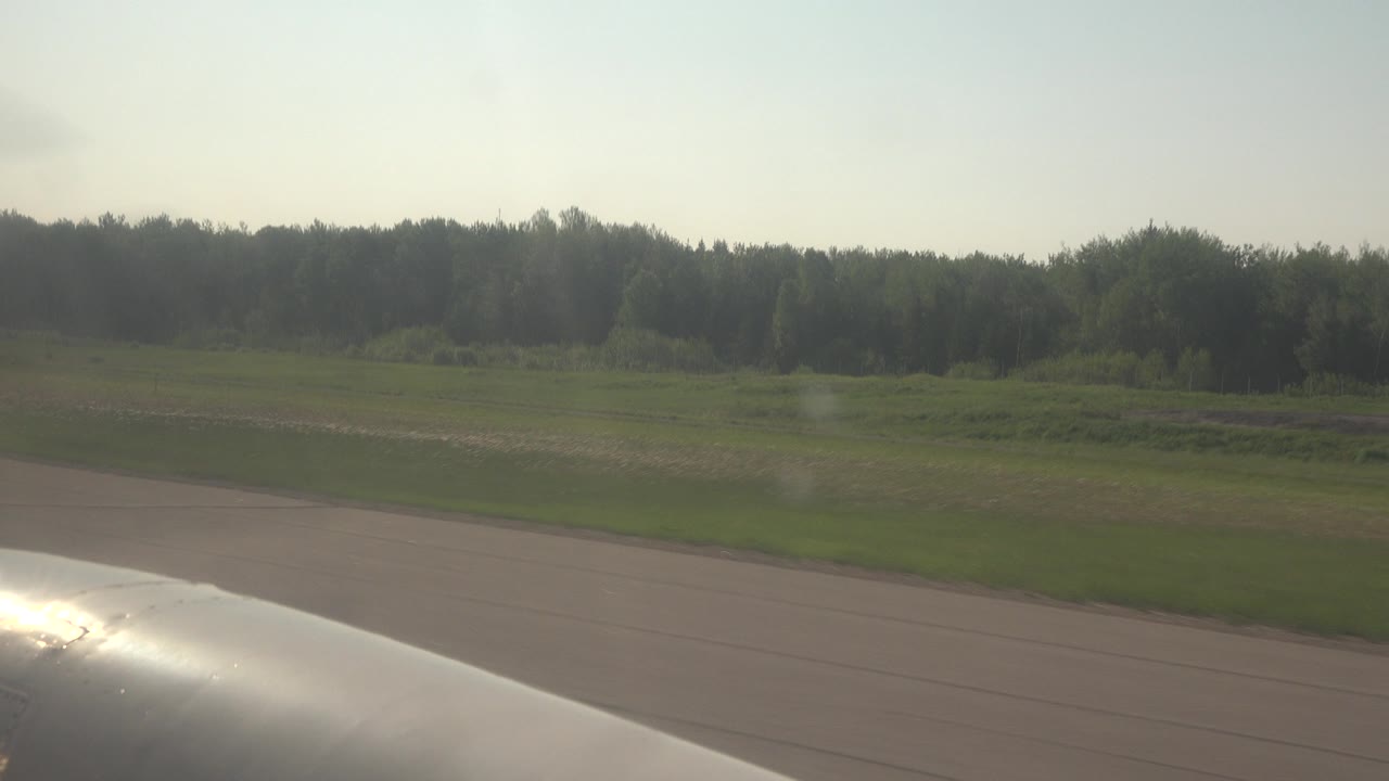 Opening frame of a Red Lake, Canada car-window view with a bright silver vehicle edge at the lower left, pavement and green fields beyond, distant trees, hazy sky, and no visible people.