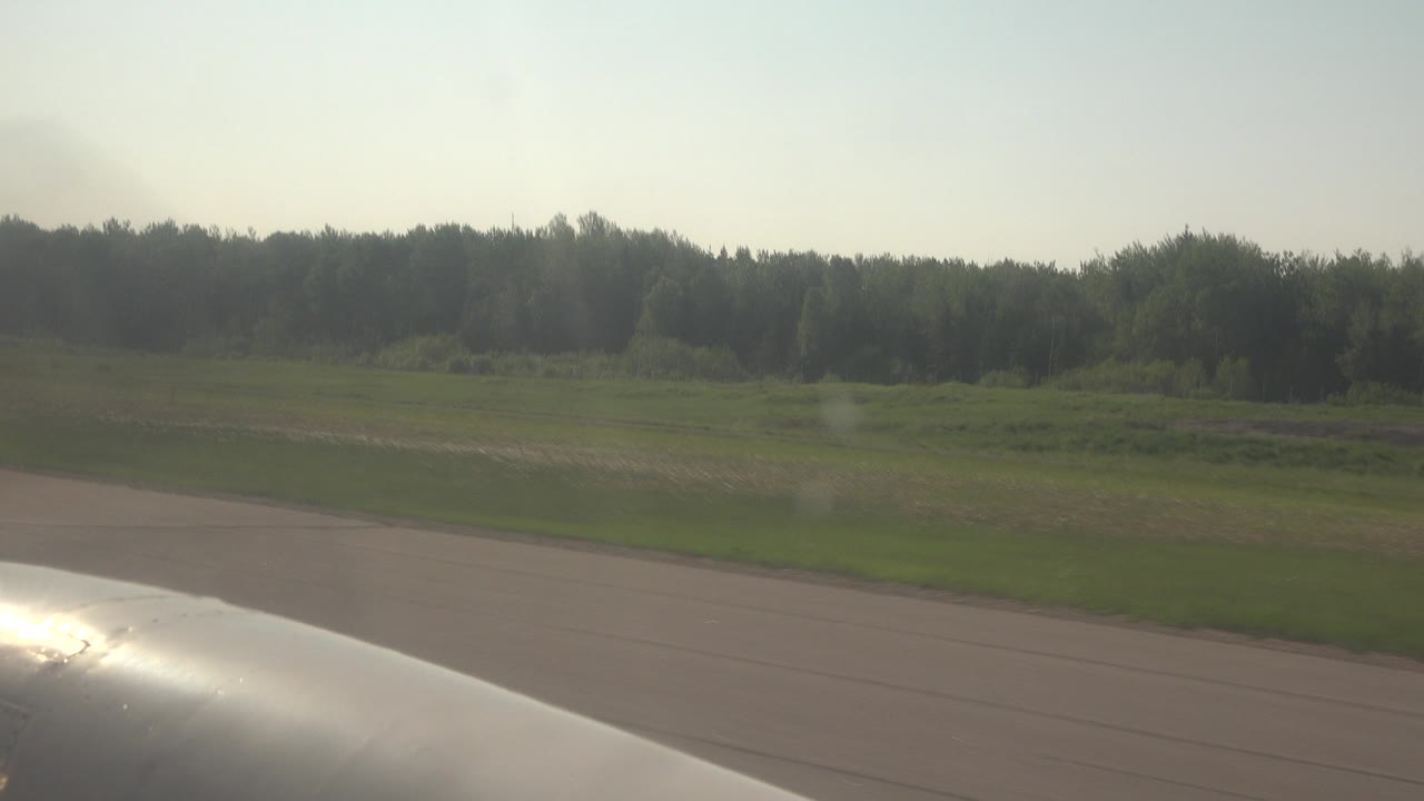 Red Lake, Canada roadside landscape seen past a silver foreground edge, with broad tan pavement, green fields, a dark tree line, pale sky, and no visible people.