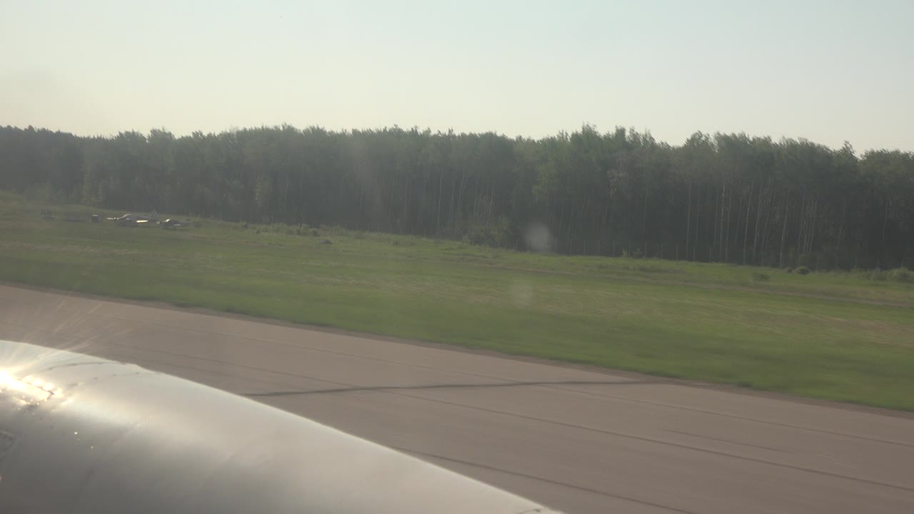 View over a reflective aircraft wing on a Red Lake, Canada runway, with grass fields, a distant tree line, and no visible people.