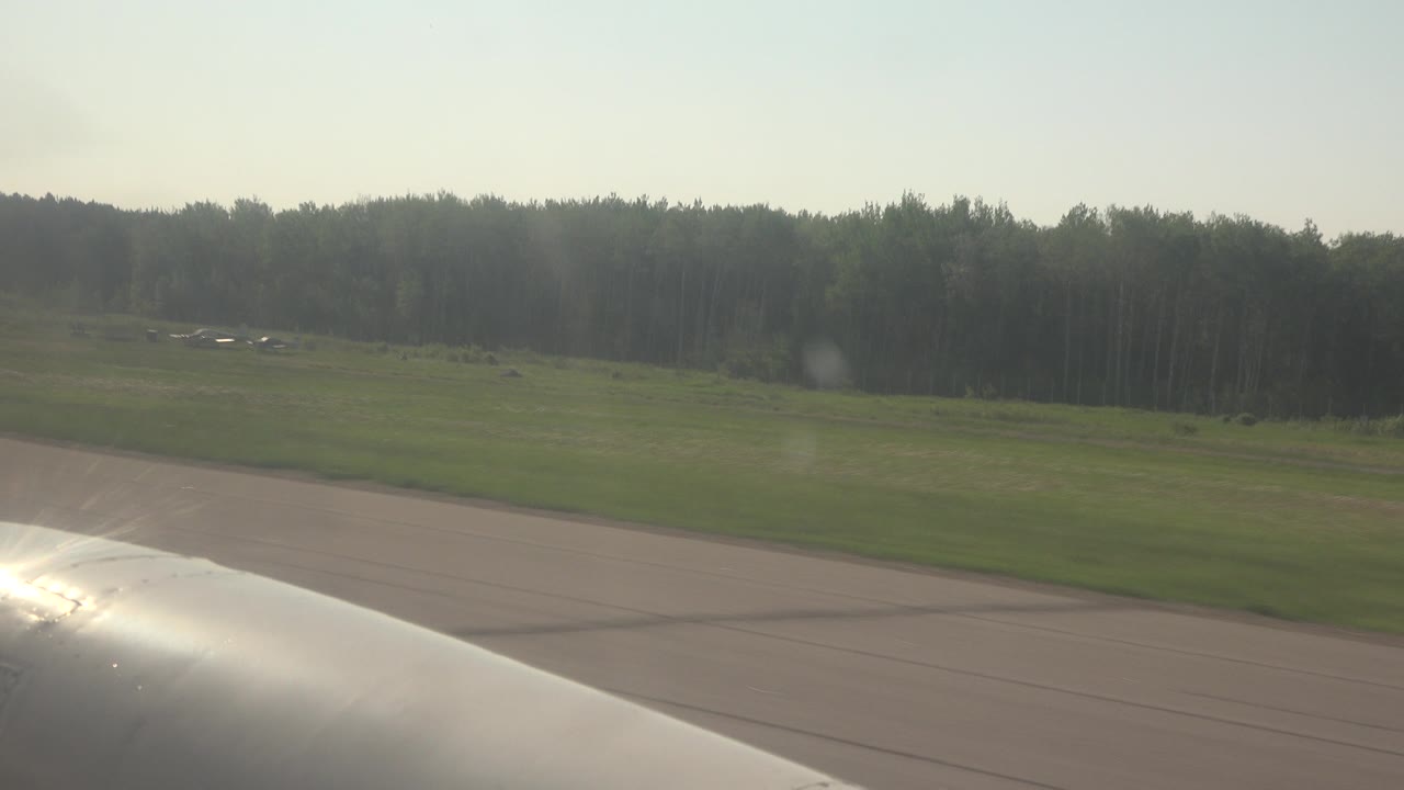 Reflective aircraft wing crosses the lower frame on a Red Lake, Canada runway, with open grass, forest in the distance, and no visible people.