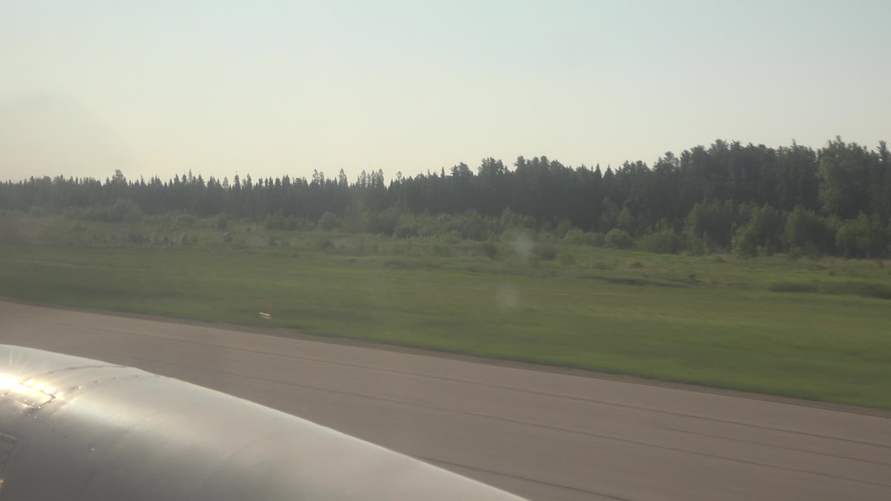 At 0:00 in Red Lake, Canada, a silver airplane wing sits low in the passenger-window view beside runway pavement, green field, and a distant forest line, with no visible people.