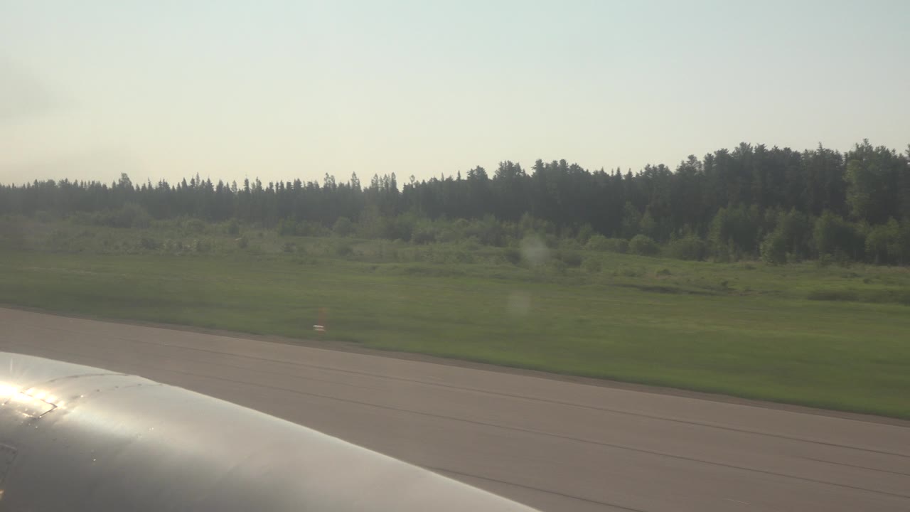 At 0:01 in Red Lake, Canada, the airplane wing reflects bright light while runway pavement and a broad green airport edge stretch toward a dark wooded horizon, with no visible people.