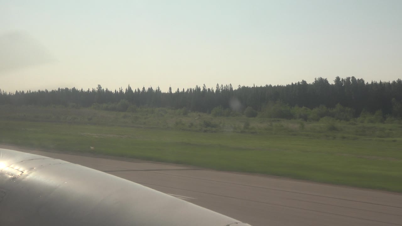 Airplane wing with runway, field, and forest in background.