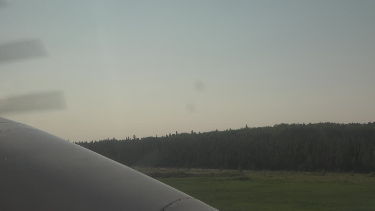 At 0:00 in Red Lake, Canada, a plane surface fills the lower foreground while blurred propeller bands cross the pale sky above a green field and forest, with no visible people.