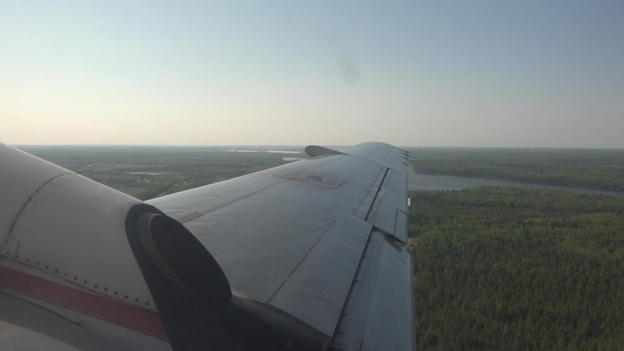 The airplane wing remains prominent over Red Lake, Canada, with dark green trees below, a low horizon, hazy clear-day light, and no visible people.