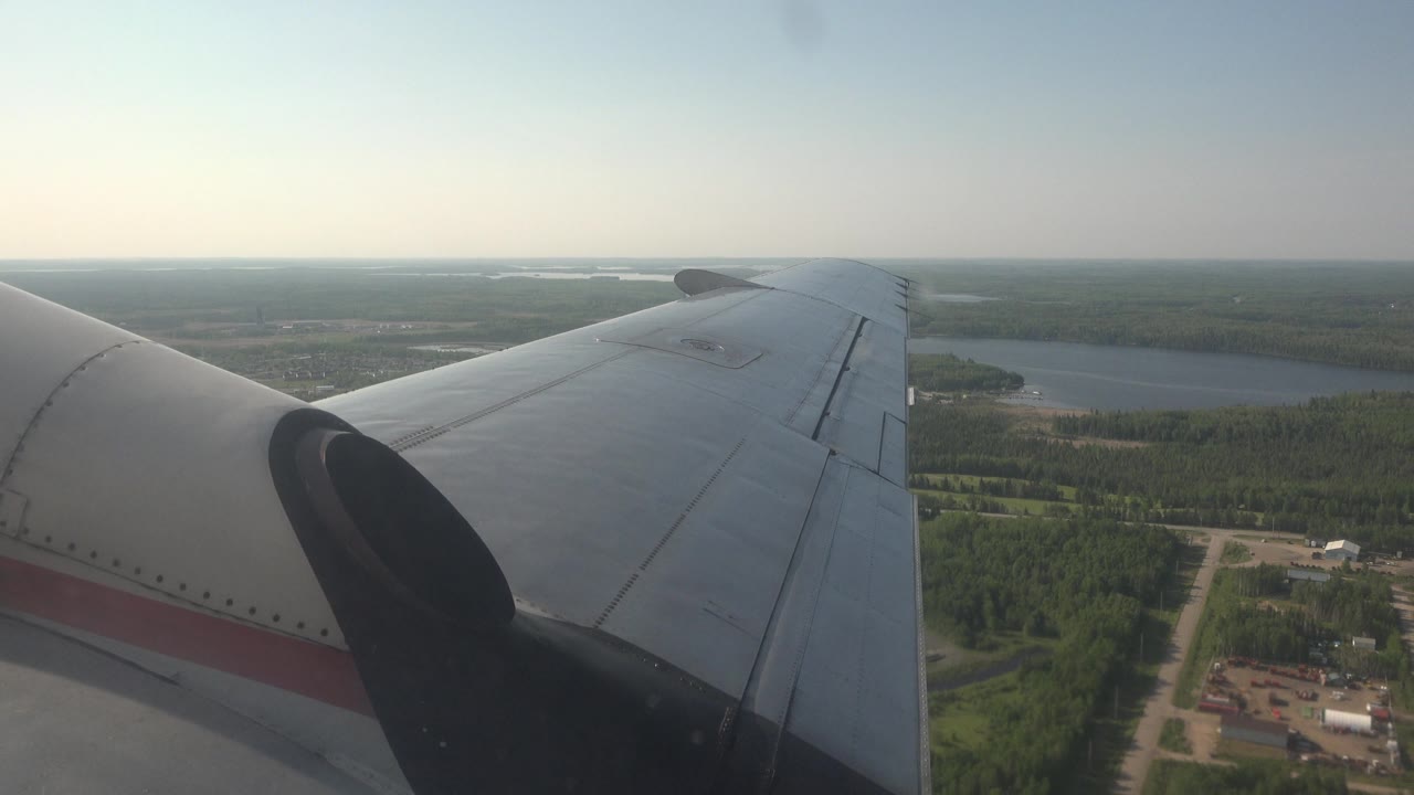 Aircraft wing and dark engine nacelle fill the foreground above green forest and a blue lake near Red Lake, Canada, with no visible people.