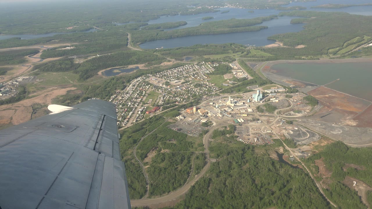 Aerial view over Red Lake, Canada, showing the aircraft wing, a dense patch of homes, curving roads, light-colored industrial structures, green forest, water, and no visible people.