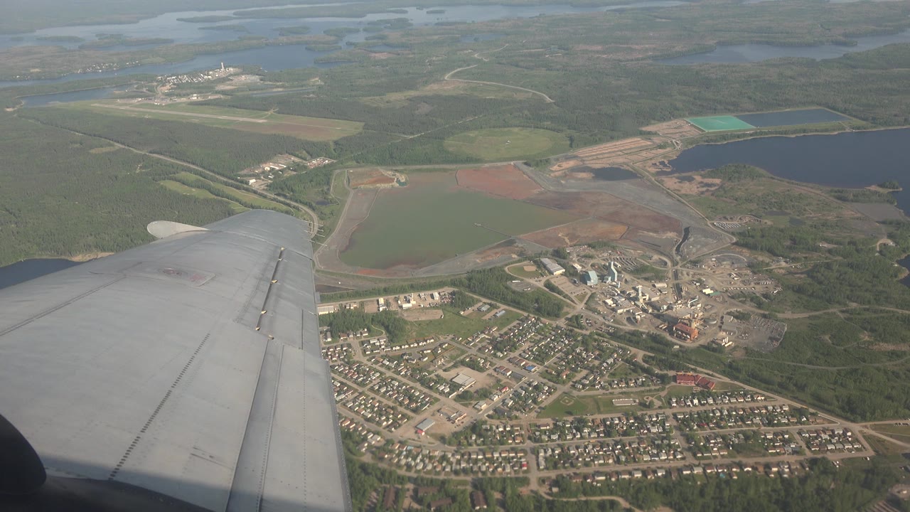 At 0:01, the wing remains in the lower frame as Red Lake's roads, homes, pale work areas, and nearby dark water sit under hazy daylight with no visible people.