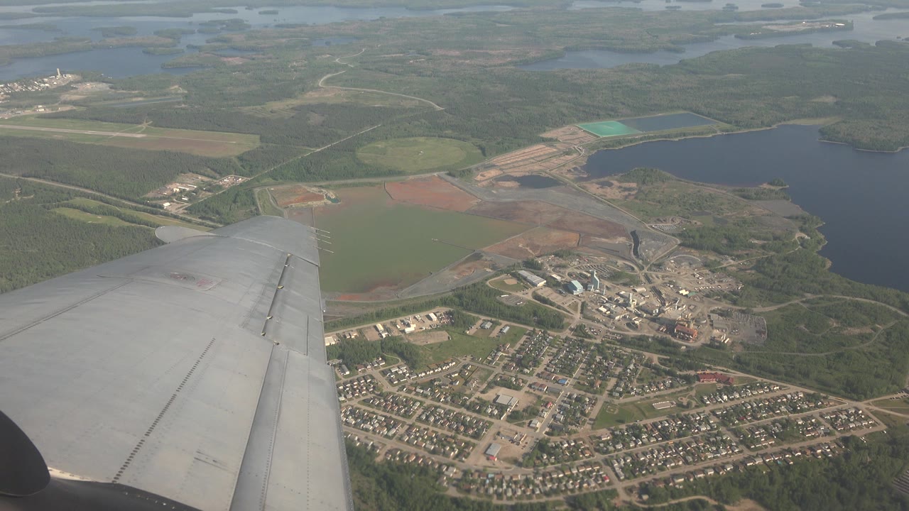 Opening frame from Red Lake, Canada showing a gray airplane wing in the lower left foreground above lakes, forest, fields, roads, and a small settlement, with no visible people.