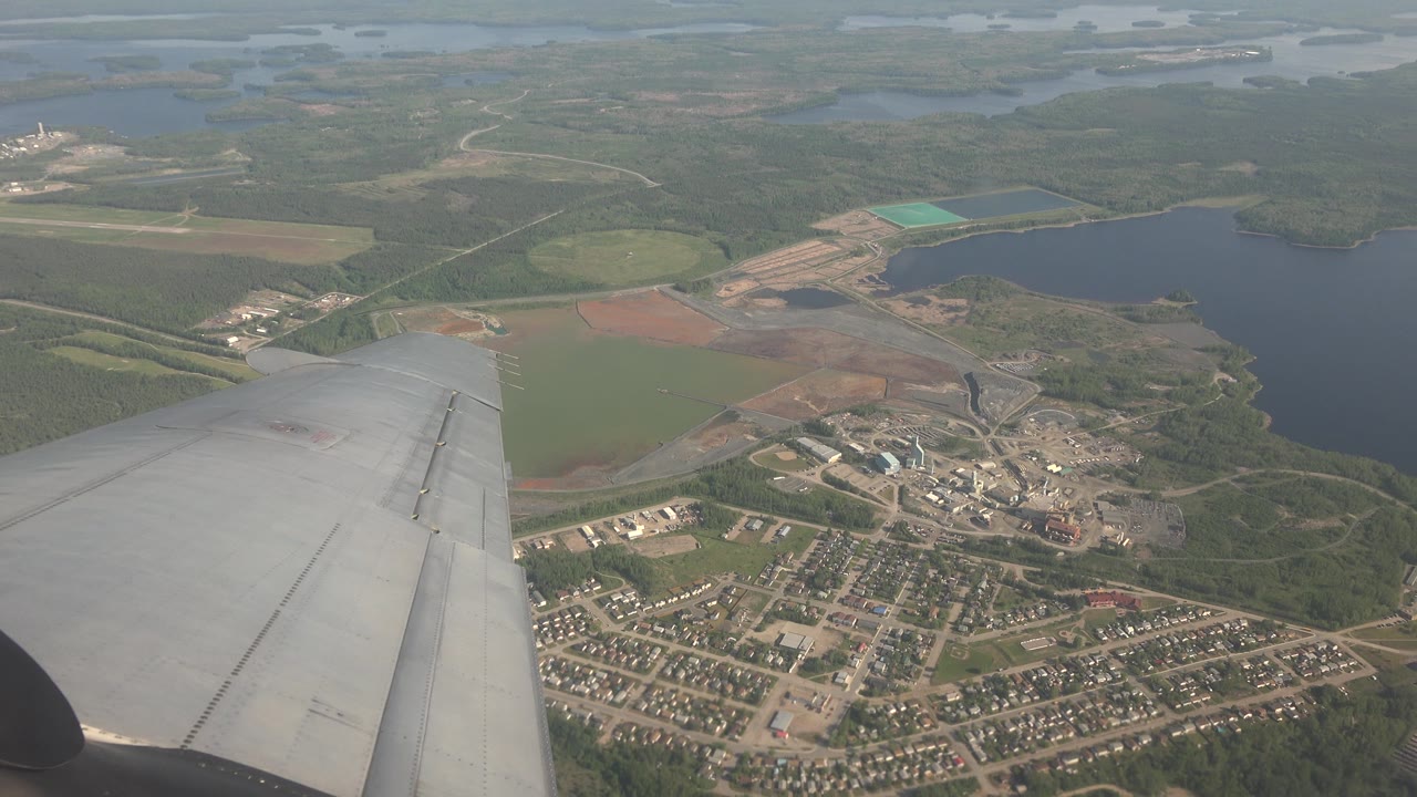 Airplane wing over a lake, forest, fields, and scattered buildings.