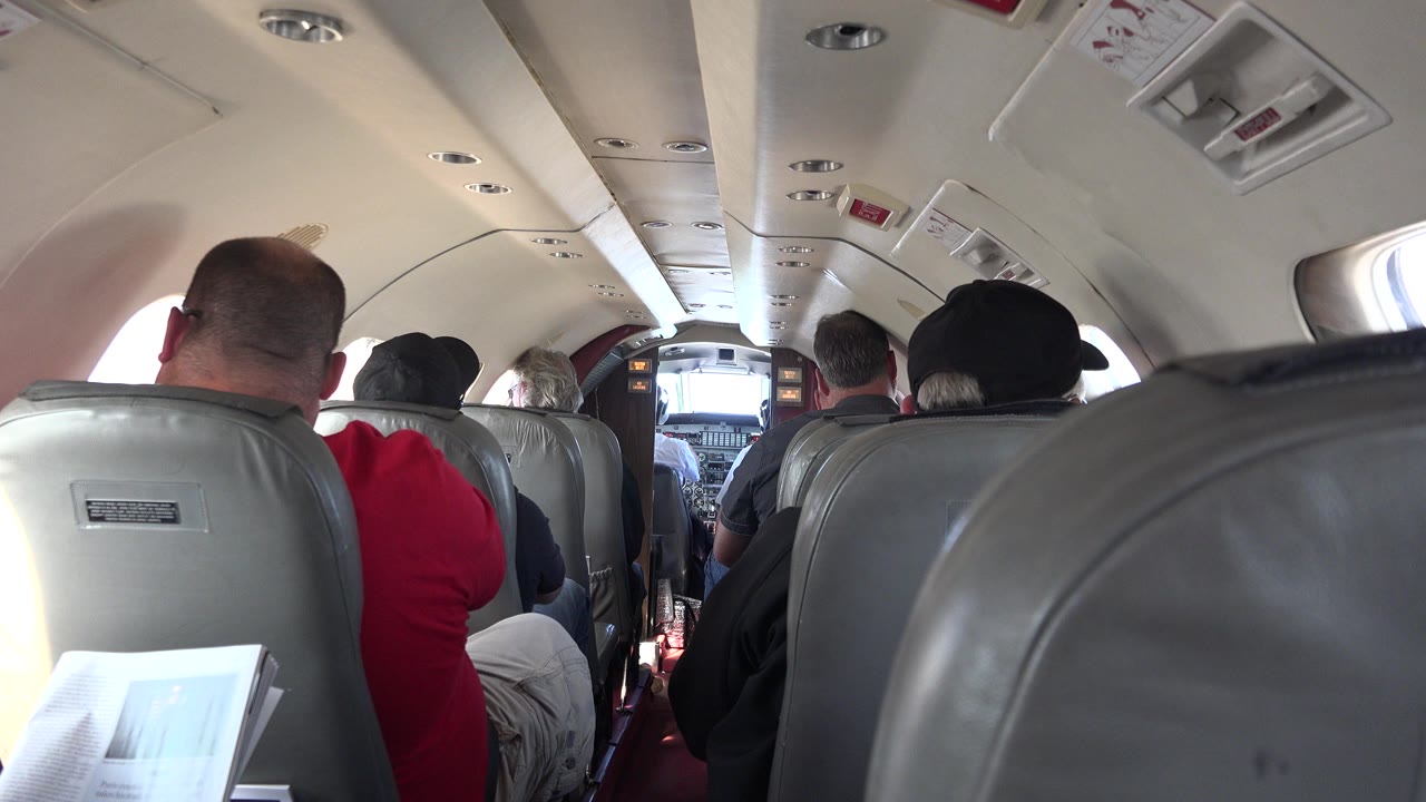 Inside an airplane cabin in Red Lake, Canada footage, the camera looks past gray seatbacks toward passengers seated ahead and a bright cockpit doorway.