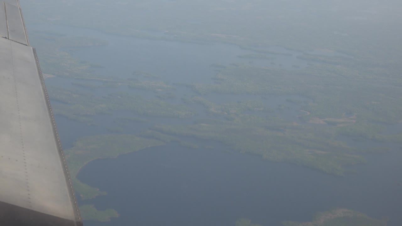 A pale airplane wing cuts down the left side over broad blue water and marshy land patches in Red Lake, Canada, with no visible people.