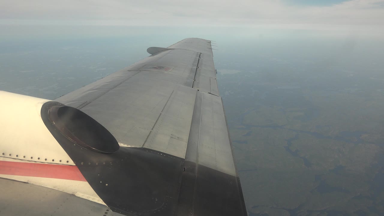Airplane wing and cream fuselage edge over hazy Red Lake, Canada terrain at the start of the clip, with no visible people.