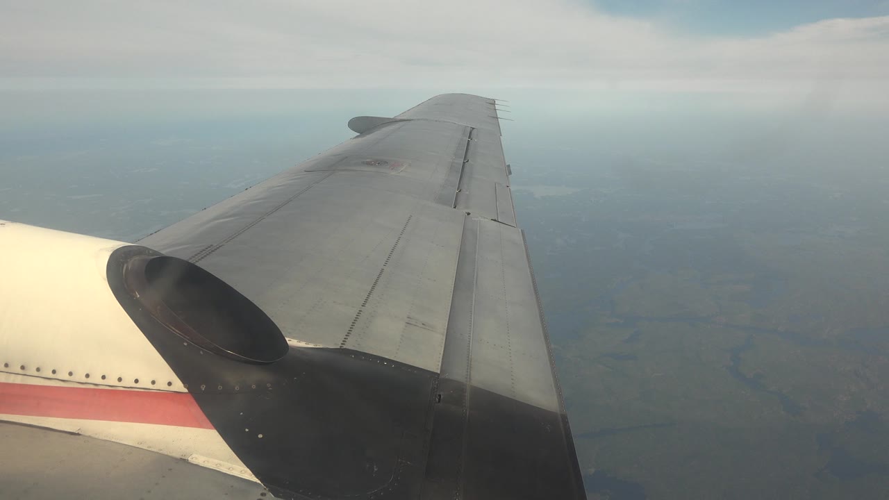 At 0:00, a gray aircraft wing and cream fuselage with a red stripe fill the left foreground over hazy Red Lake, Canada terrain, with no visible people.