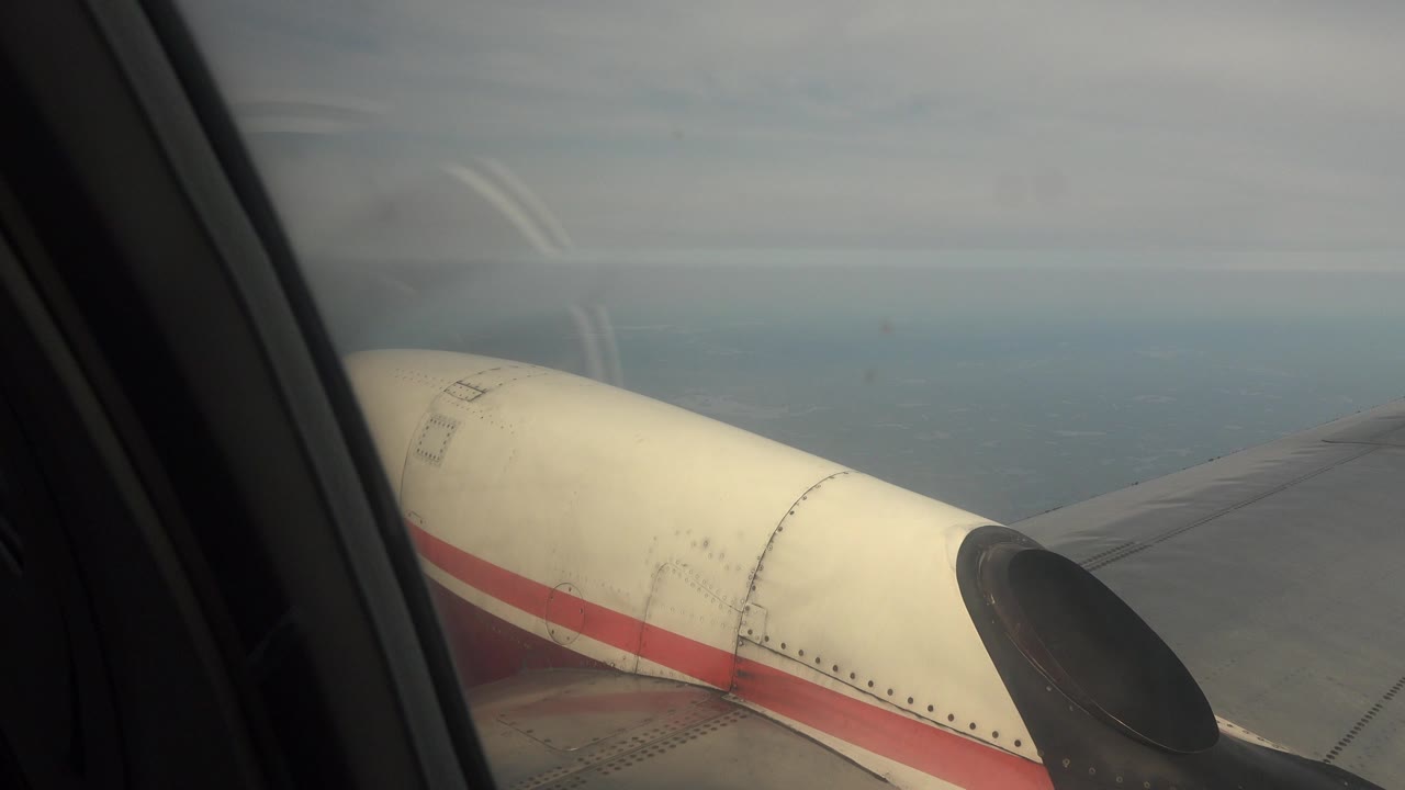Passenger-window view in Red Lake, Canada showing a cream and red airplane engine cowling, dark propeller area, wing, hazy water, and no visible people at the start of the clip.