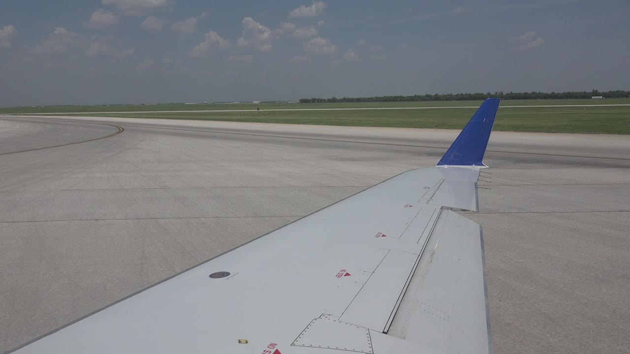 White airplane wing with a blue winglet seen over Winnipeg runway pavement and grass under scattered clouds, with no visible people.