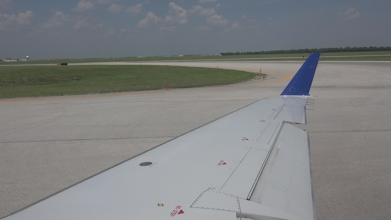 Start of the Winnipeg, Canada clip showing a white airplane wing with a blue wingtip over pale taxiway concrete, green grass, distant runway, and no visible people.
