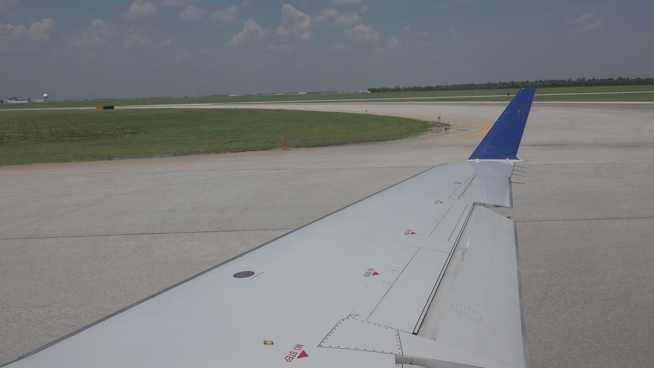 One second in, the blue-tipped airplane wing remains in the foreground while the Winnipeg airport pavement and grassy field extend across the background with no visible people.