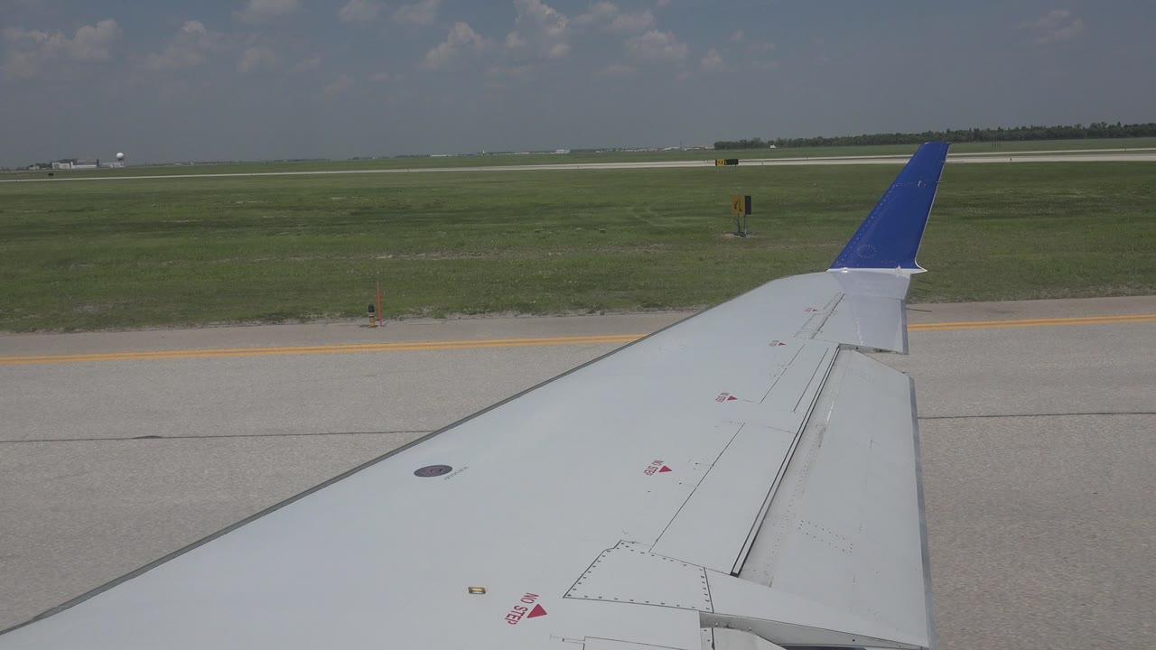 Winnipeg runway view from a passenger window, showing the blue-tipped wing angled across gray pavement, a green airfield, low horizon, clouds, and no visible people.