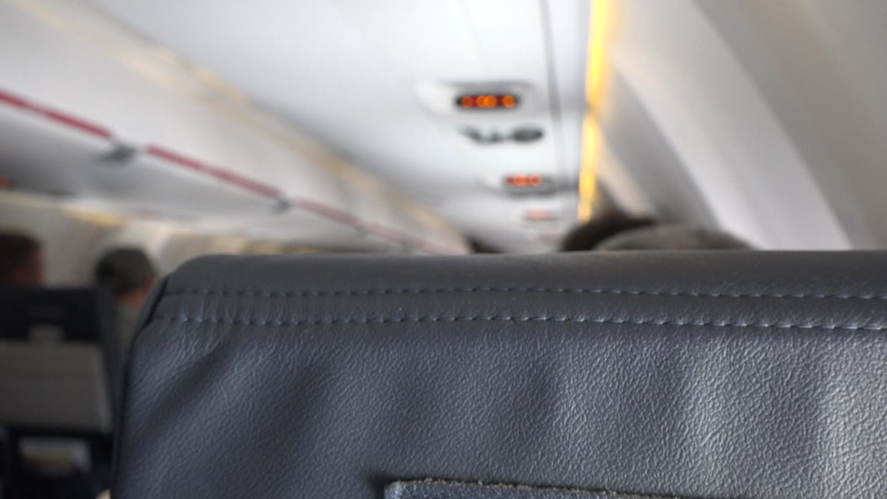 The cabin remains framed over the top of a dark seatback, with the ceiling panels and small amber overhead indicators visible in the background.