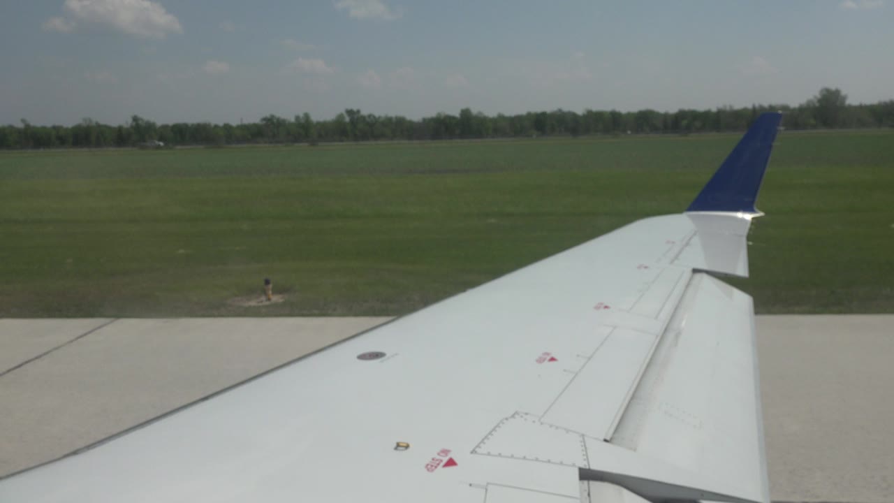 White airplane wing with a blue tip seen from a passenger window in Winnipeg, Canada, with concrete runway, green grass, distant trees, and no visible people.