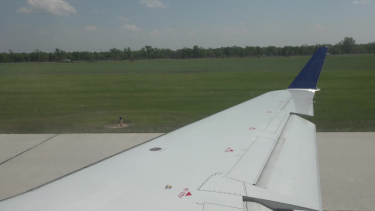 Blue-tipped wing remains centered over runway concrete in Winnipeg, Canada, with open grass fields and a low horizon under daylight, no visible people.