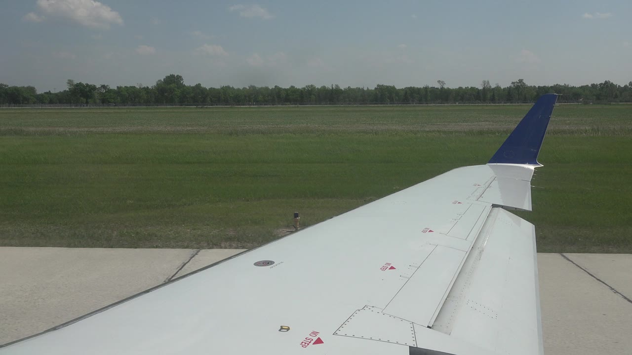 The airplane wing remains low in frame as the grassy field and distant tree line stretch across the Winnipeg runway background, with no visible people.