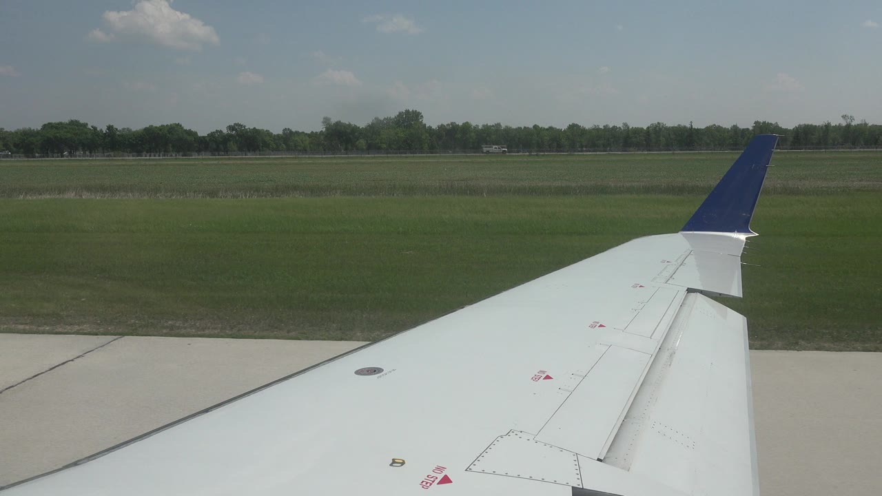 White airplane wing with a blue winglet seen over runway concrete and a green field in Winnipeg, Canada, with no visible people.