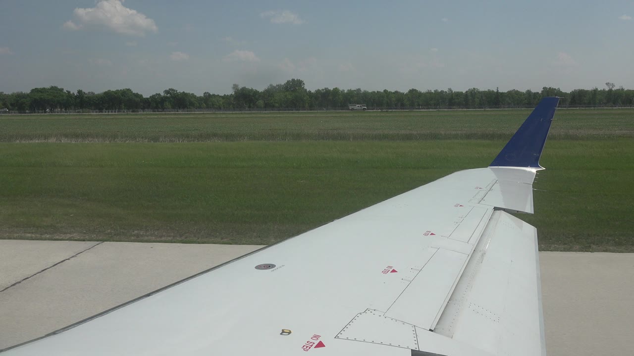 Airplane wing remains steady in the foreground while runway pavement, a broad green field, and distant trees fill the Winnipeg, Canada background, with no visible people.