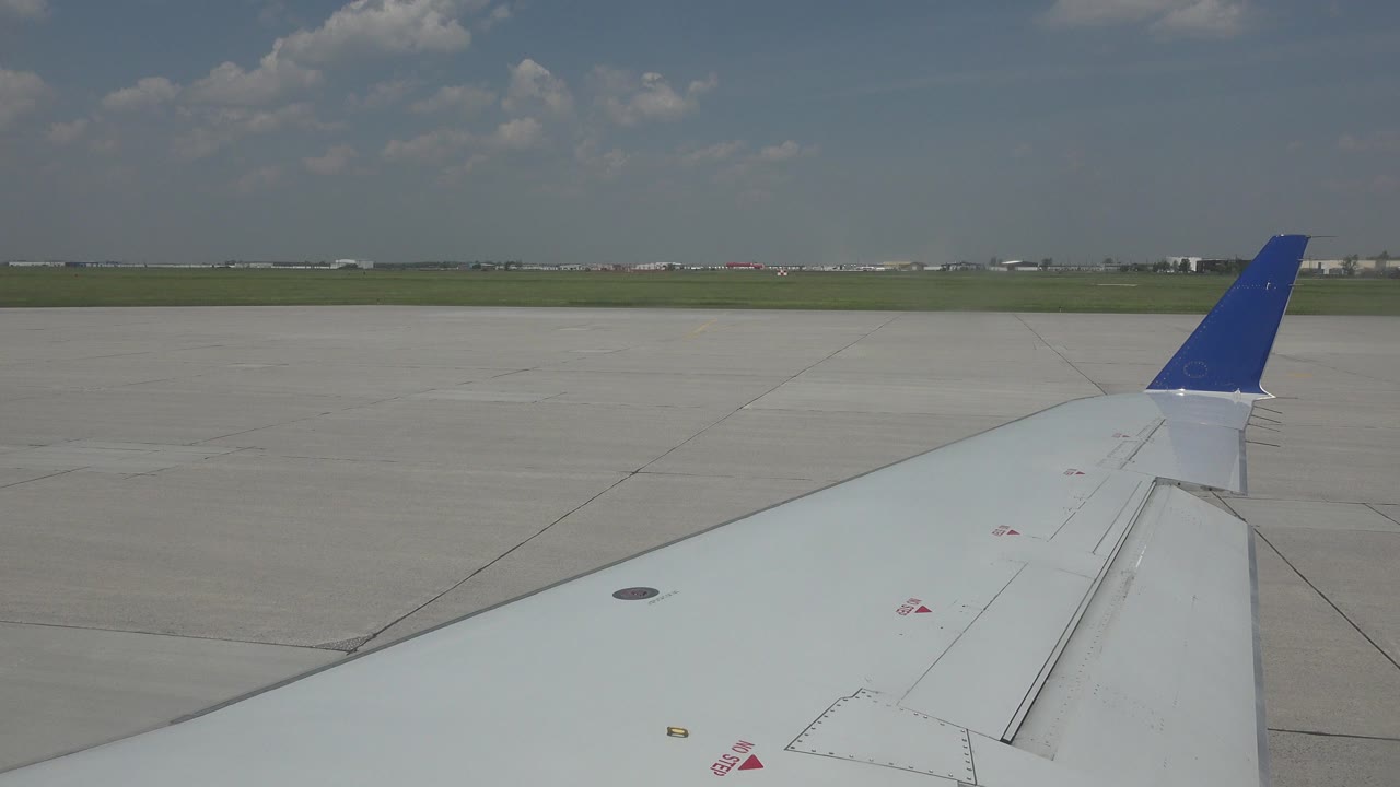 Passenger-window view in Winnipeg, Canada, showing a white airplane wing with a blue winglet over pale airport pavement and a low green horizon, with no visible people.