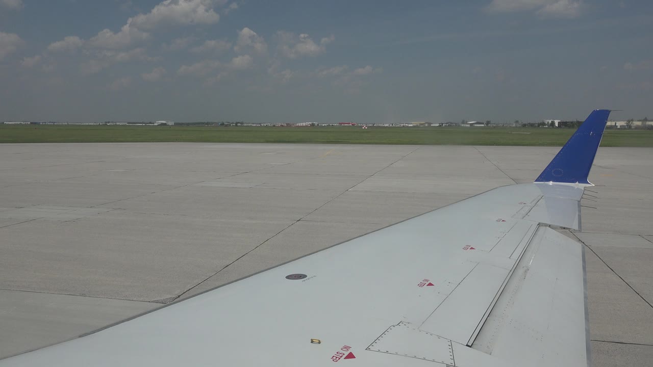 The wing remains prominent from the passenger-window angle, with its blue winglet near the horizon and broad concrete airport pavement stretching across the frame, with no visible people.