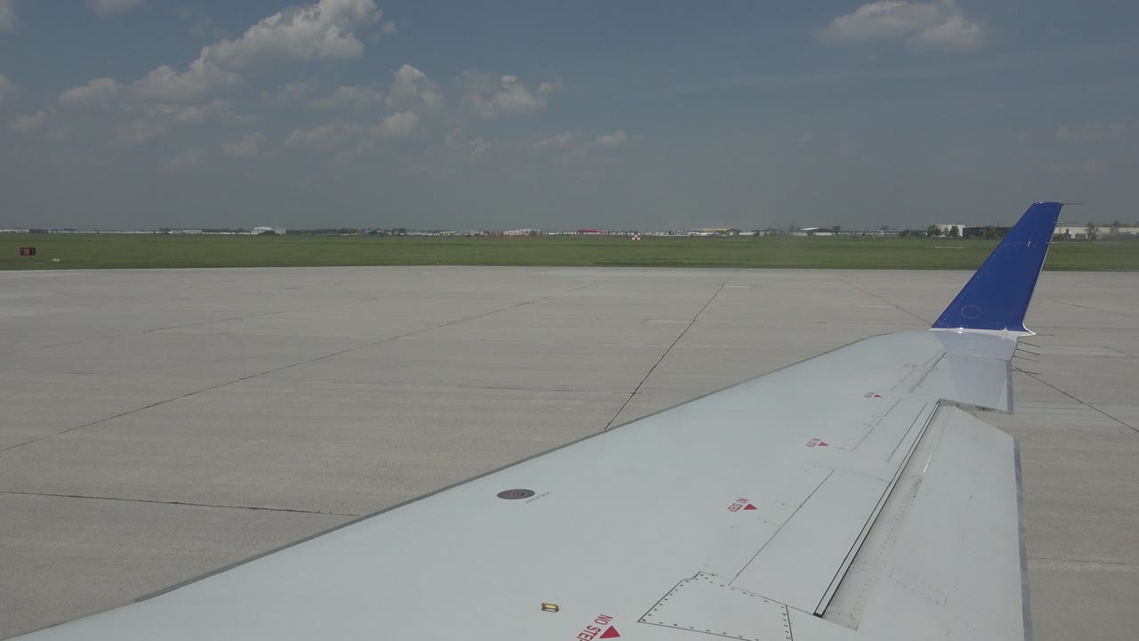 Passenger-window view of a white airplane wing and blue winglet over Winnipeg airport tarmac, with grass and distant airport structures on the horizon and no visible people.