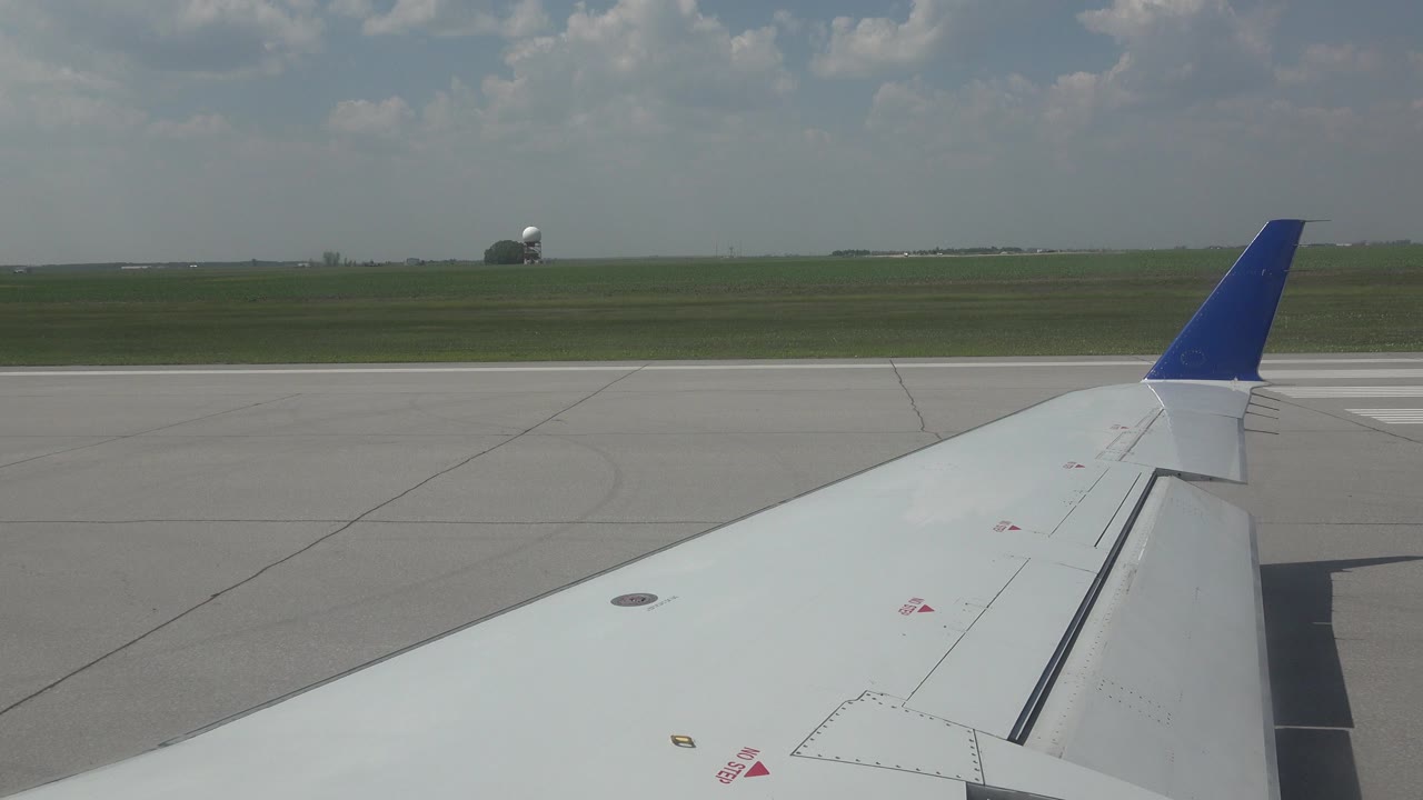 Passenger-window view near Winnipeg, Canada, showing a white airplane wing with blue winglet over gray runway pavement, green field beyond, cloudy sky, and no visible people.